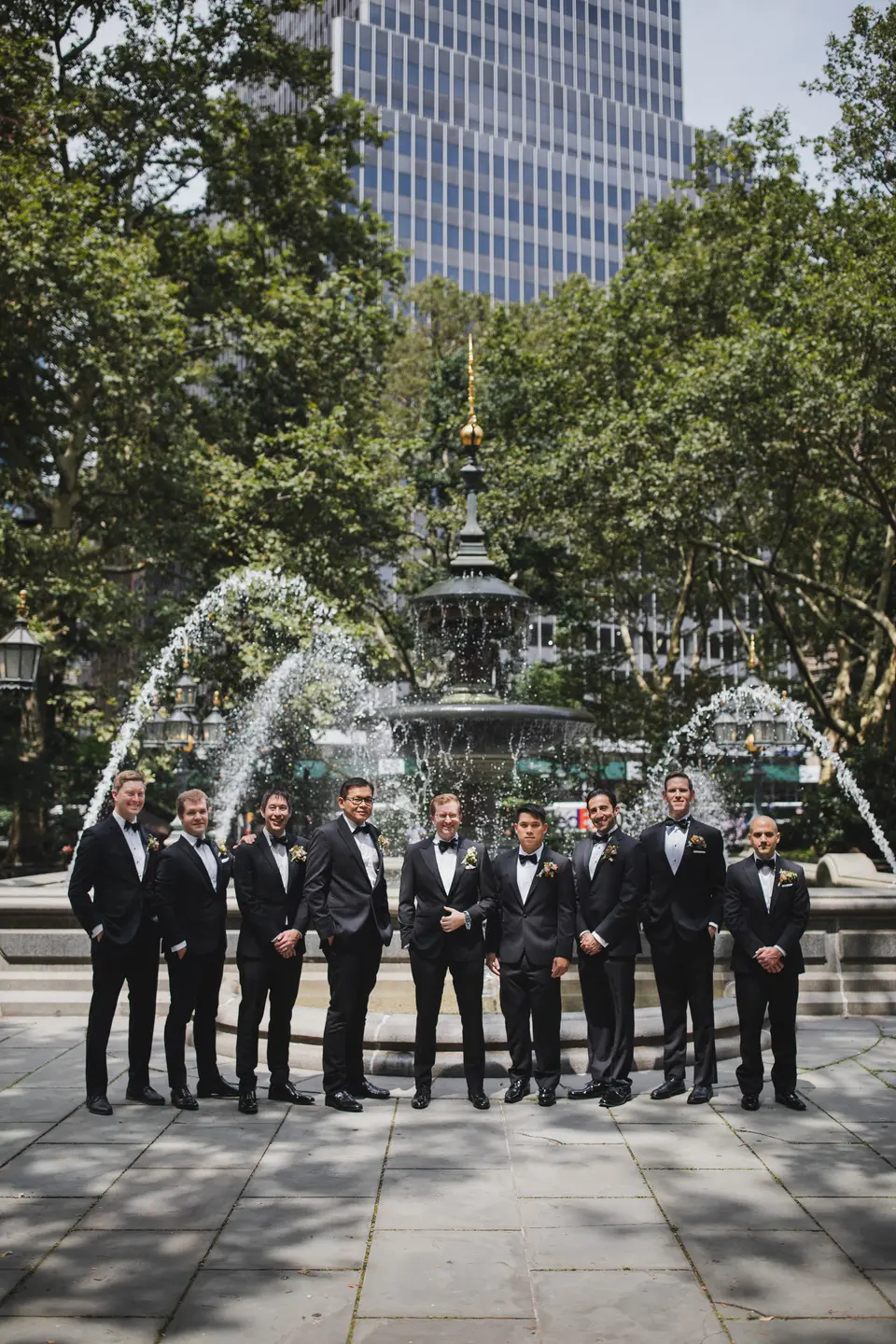 The groomsmen stand before a fountain in a city plaza, towers rising behind the trees.