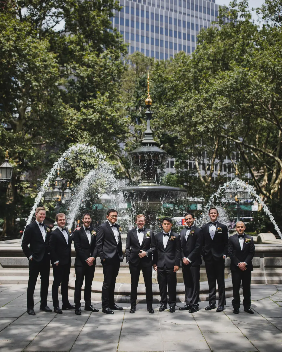 The groomsmen stand before a fountain in a city plaza, towers rising behind the trees.