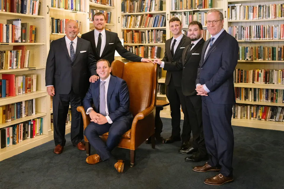 The groom sits in a leather wing chair in the club library, his groomsmen standing around him among the bookshelves.