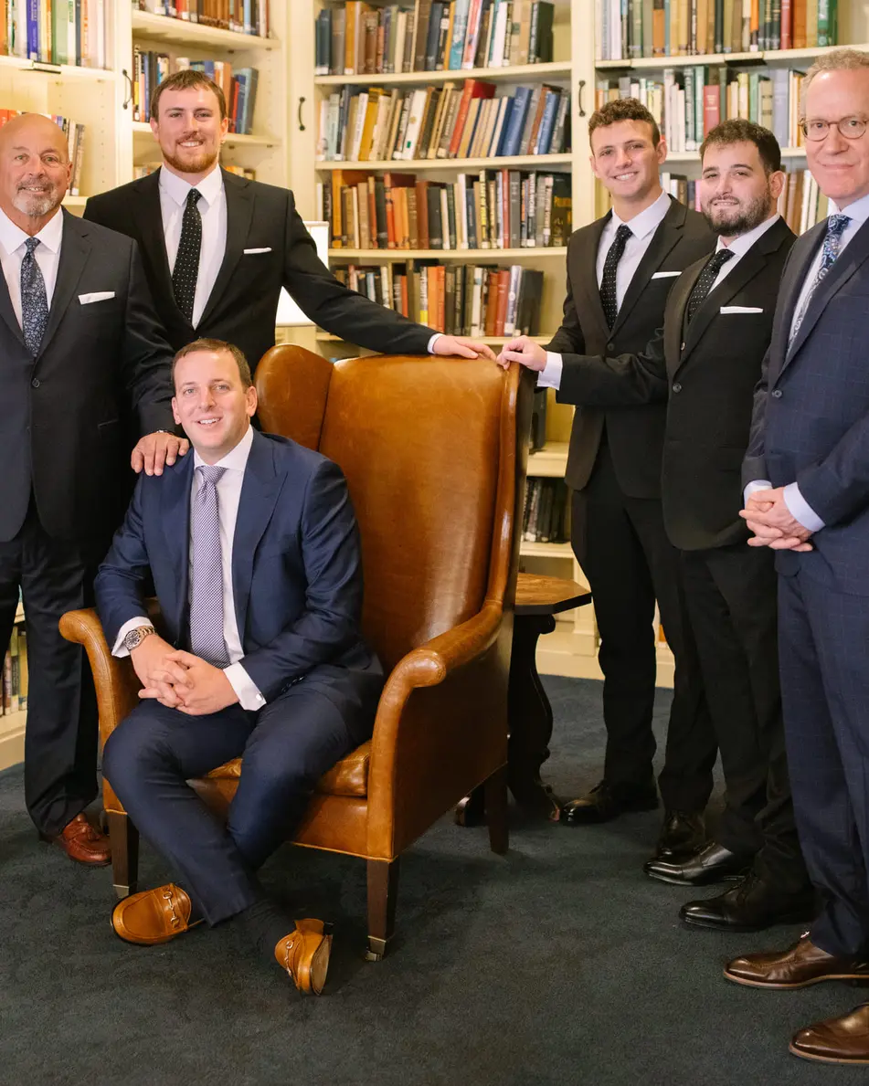 The groom sits in a leather wing chair in the club library, his groomsmen standing around him among the bookshelves.