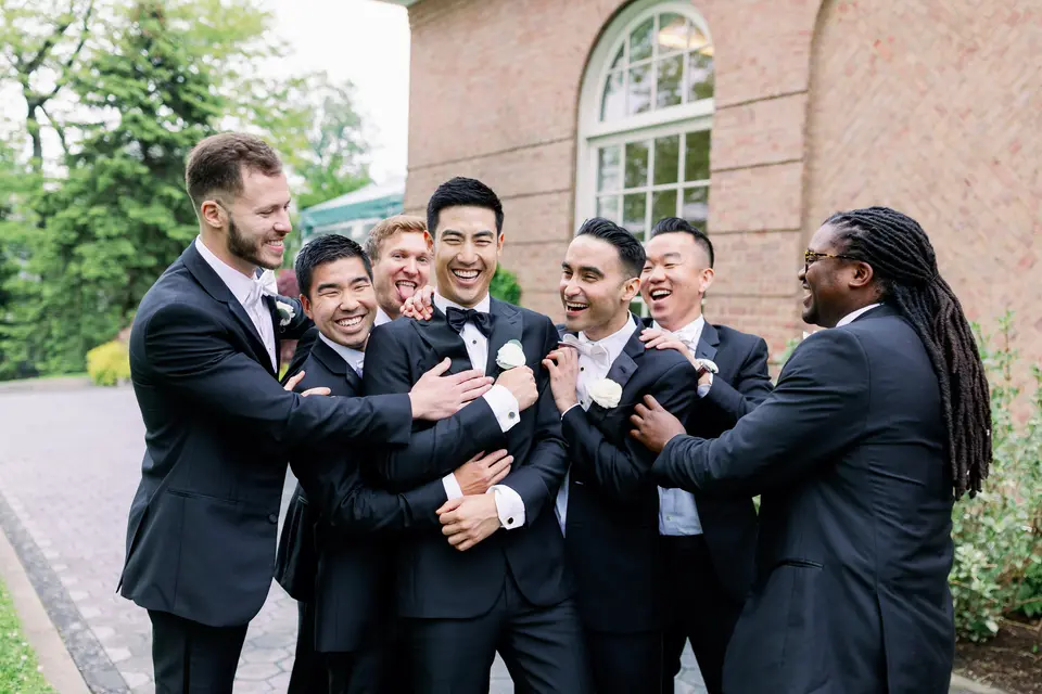 The groom's friends crowd around him in a laughing embrace outside a red brick building at the garden.