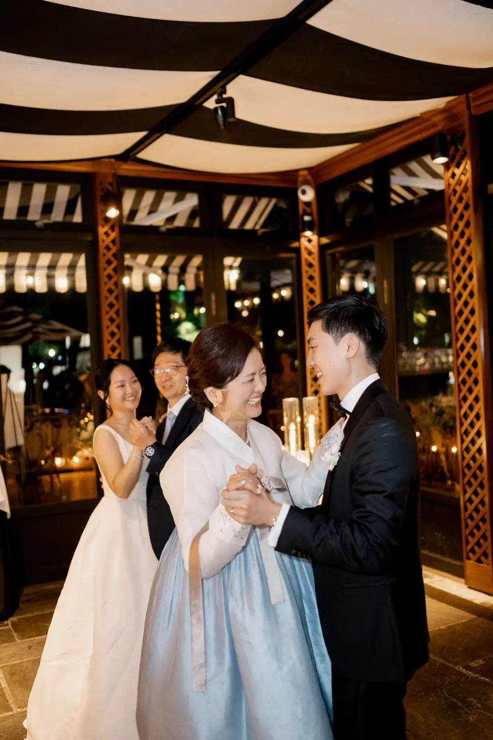 The groom dances with a woman in a pale blue hanbok beneath a striped canopy, the bride smiling beside them.