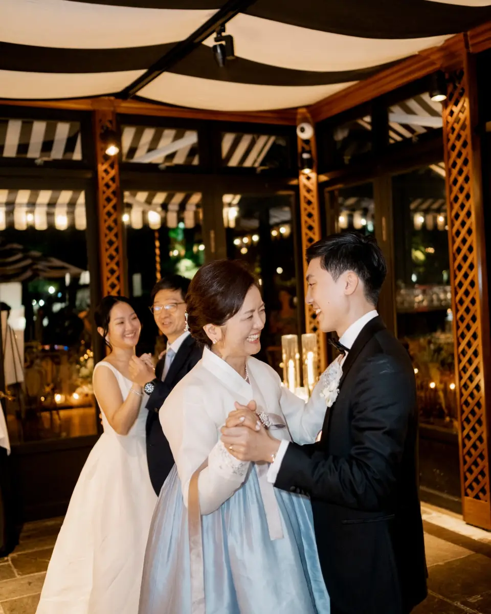 A mother in a white and pale blue hanbok laughs as she dances with the groom beneath a striped canopy, the bride and a guest smiling behind them.