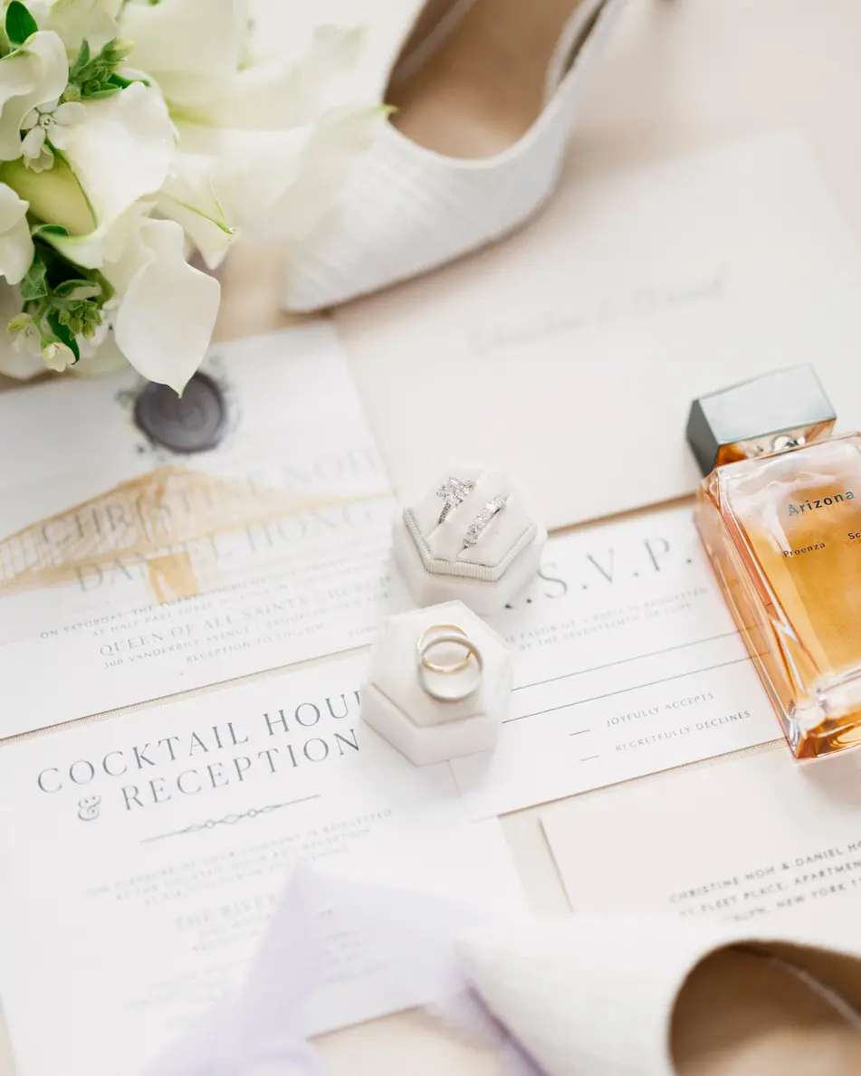 The invitation suite laid out with white flowers, pearl earrings, a bottle of perfume and the bride's shoes.