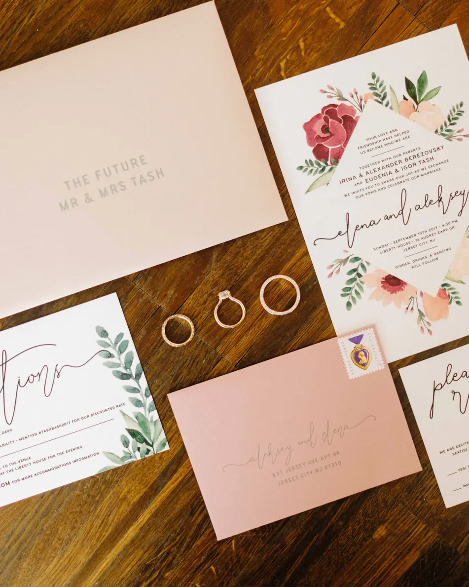 The invitation suite in blush with painted flowers, laid out with the rings on a wooden table.