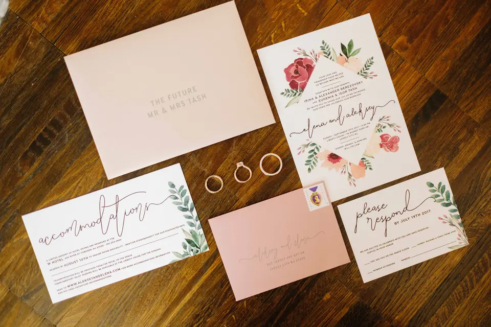 The invitation suite in blush with painted flowers, laid out with the rings on a wooden table.