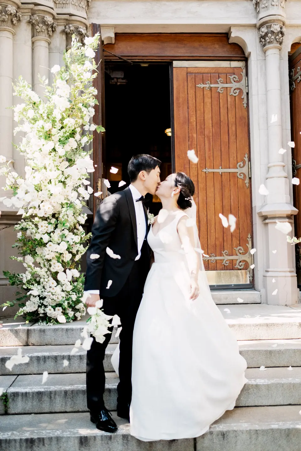 At the church door, the couple kiss as petals fall around them, white flowers climbing the stone beside them.