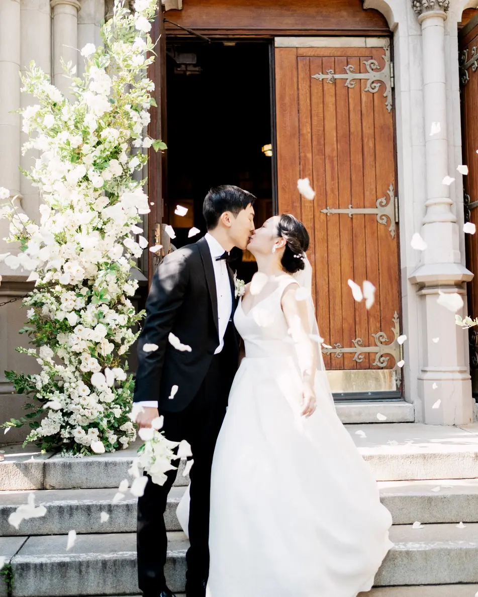At the church door, the couple kiss as petals fall around them, white flowers climbing the stone beside them.