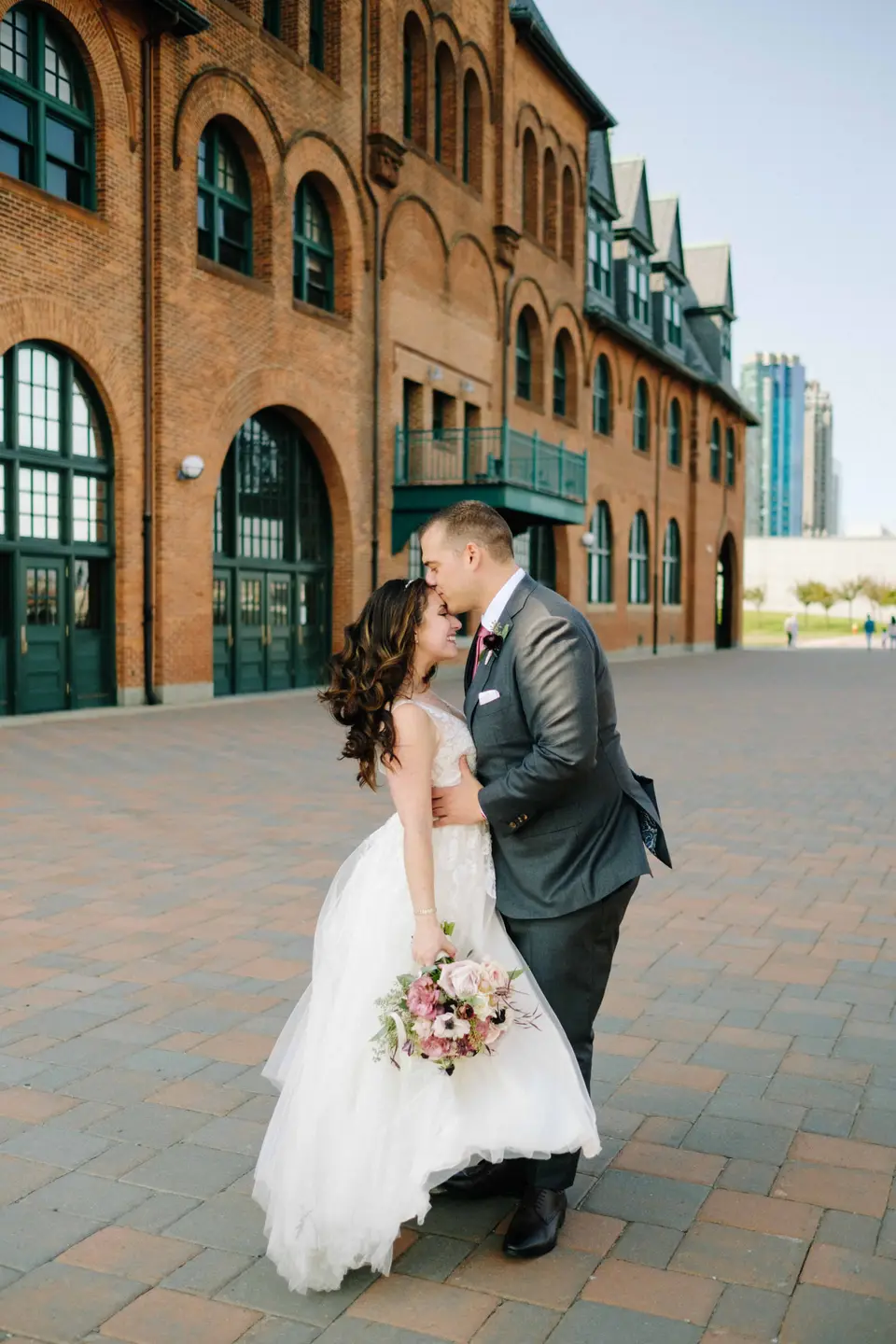 The couple kiss in front of the red brick terminal building, her bouquet of blush and burgundy flowers in hand.