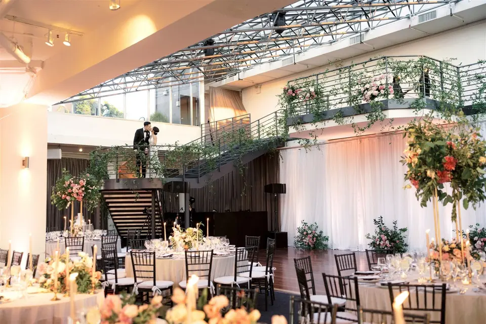 The bride and groom kiss on the black iron staircase at Tribeca Rooftop, jasmine trailing from the railings above round candlelit tables, tall blush and coral arrangements, white drapes and the wood dance floor, under the glass roof.