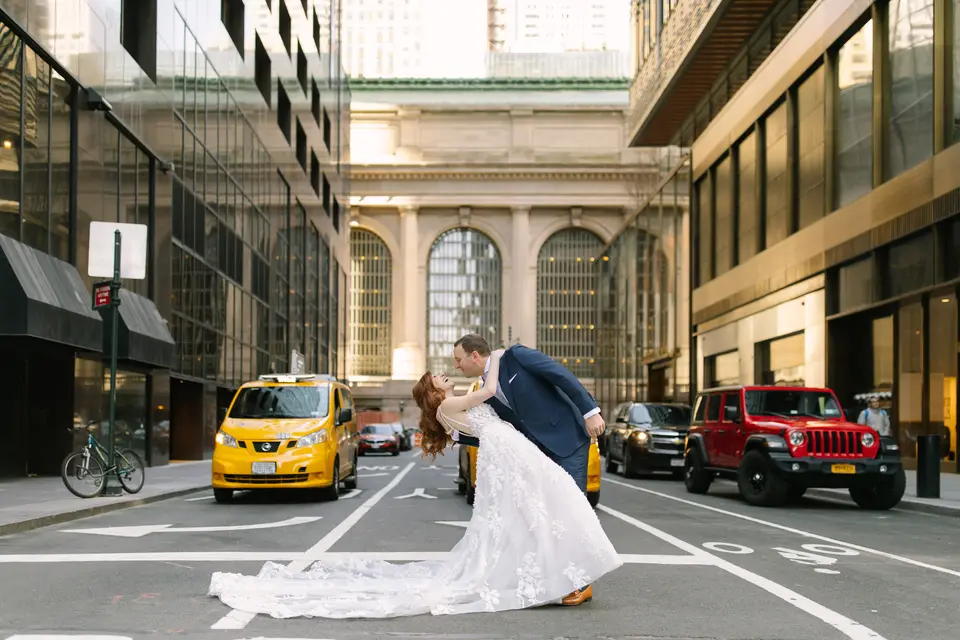 In the middle of Vanderbilt Avenue, the groom dips the bride for a kiss, Grand Central's arched windows behind them and a yellow taxi waiting.