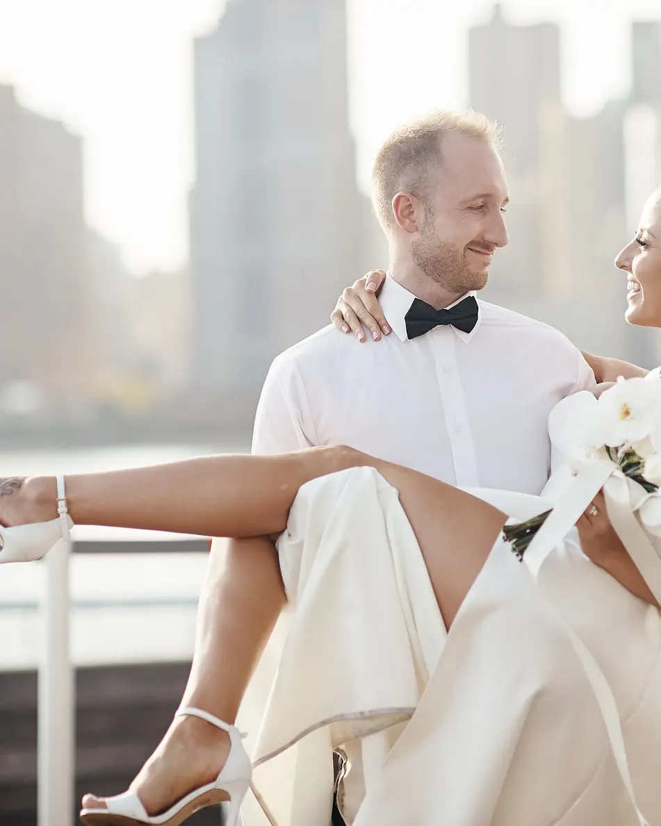 The bride sits laughing on the groom's knee by the railing, the skyline behind them.