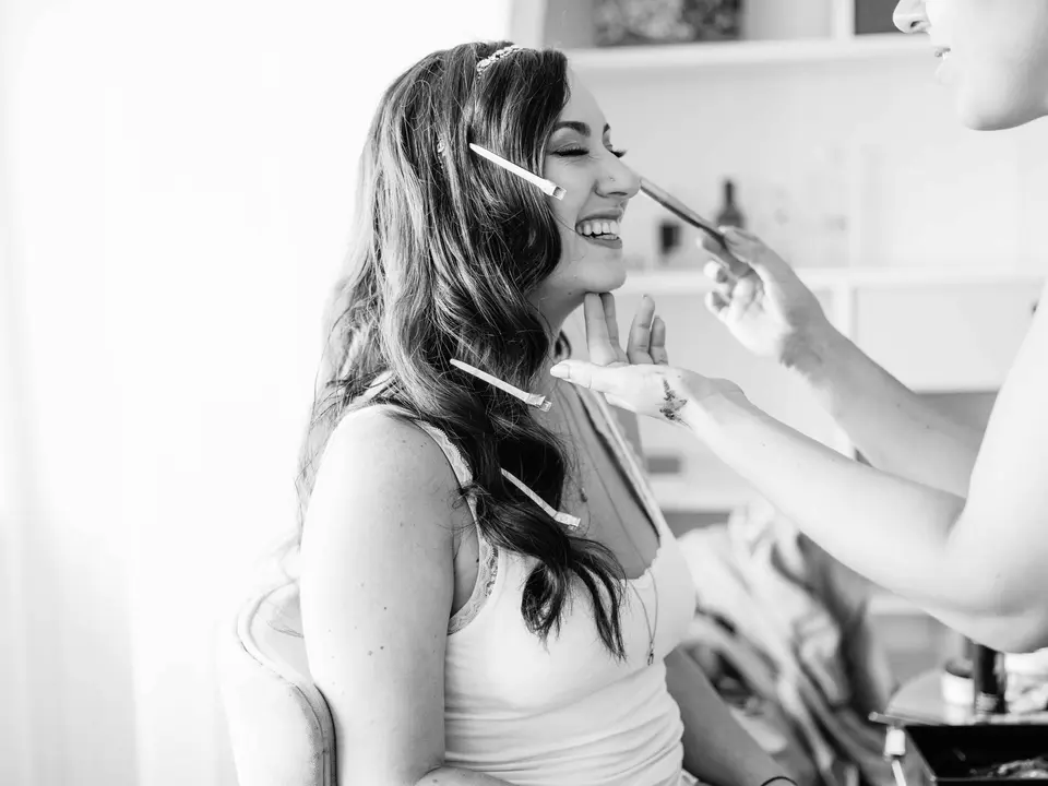 The bride laughs as her makeup is done, clips in her hair. Black and white.