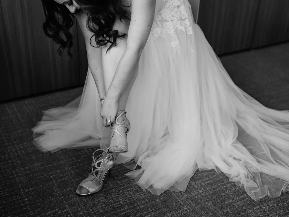The bride fastens a strappy sandal beneath the tulle of her gown. Black and white.