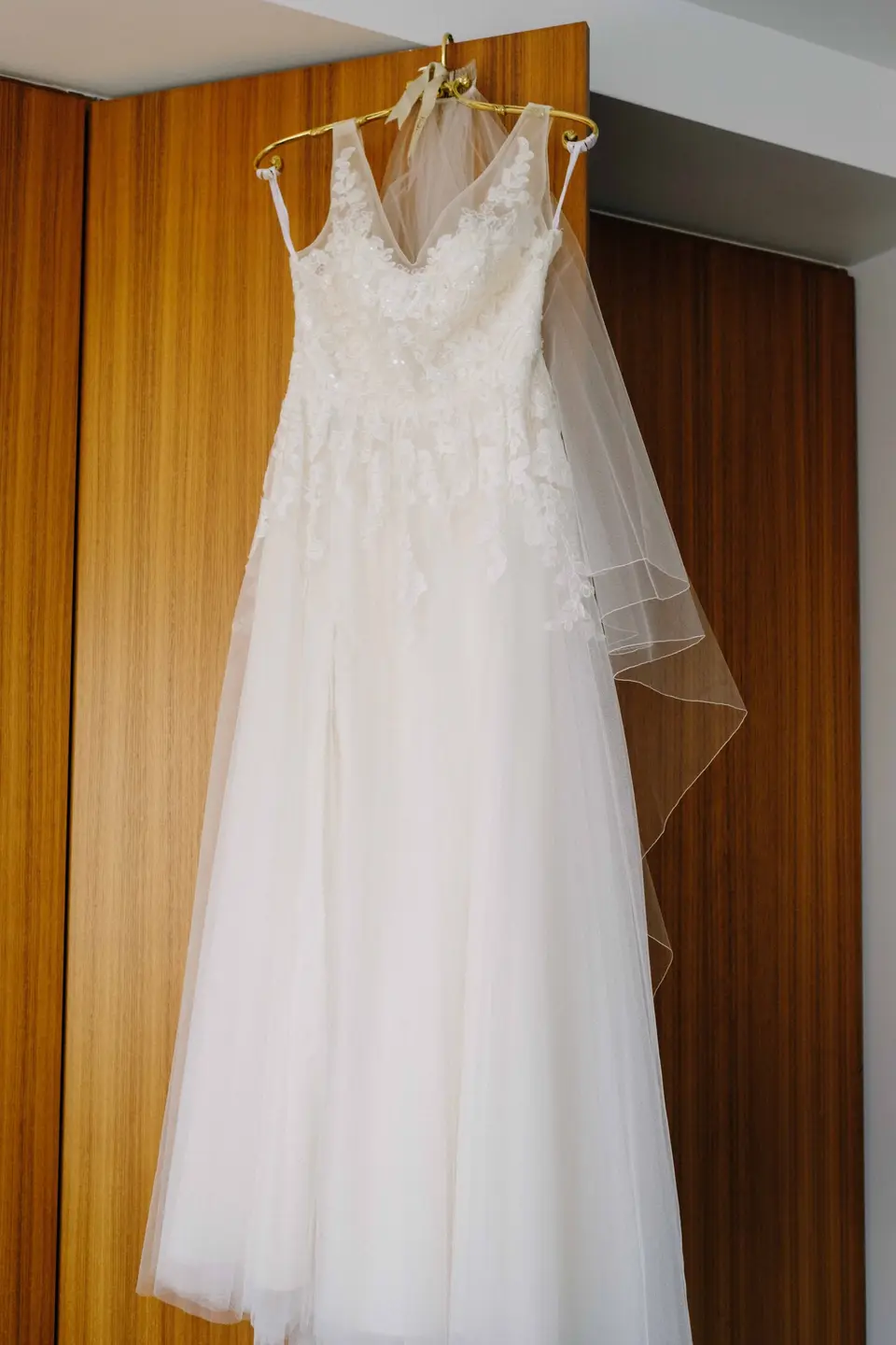 The wedding gown and veil hanging against a warm wood-panelled door.