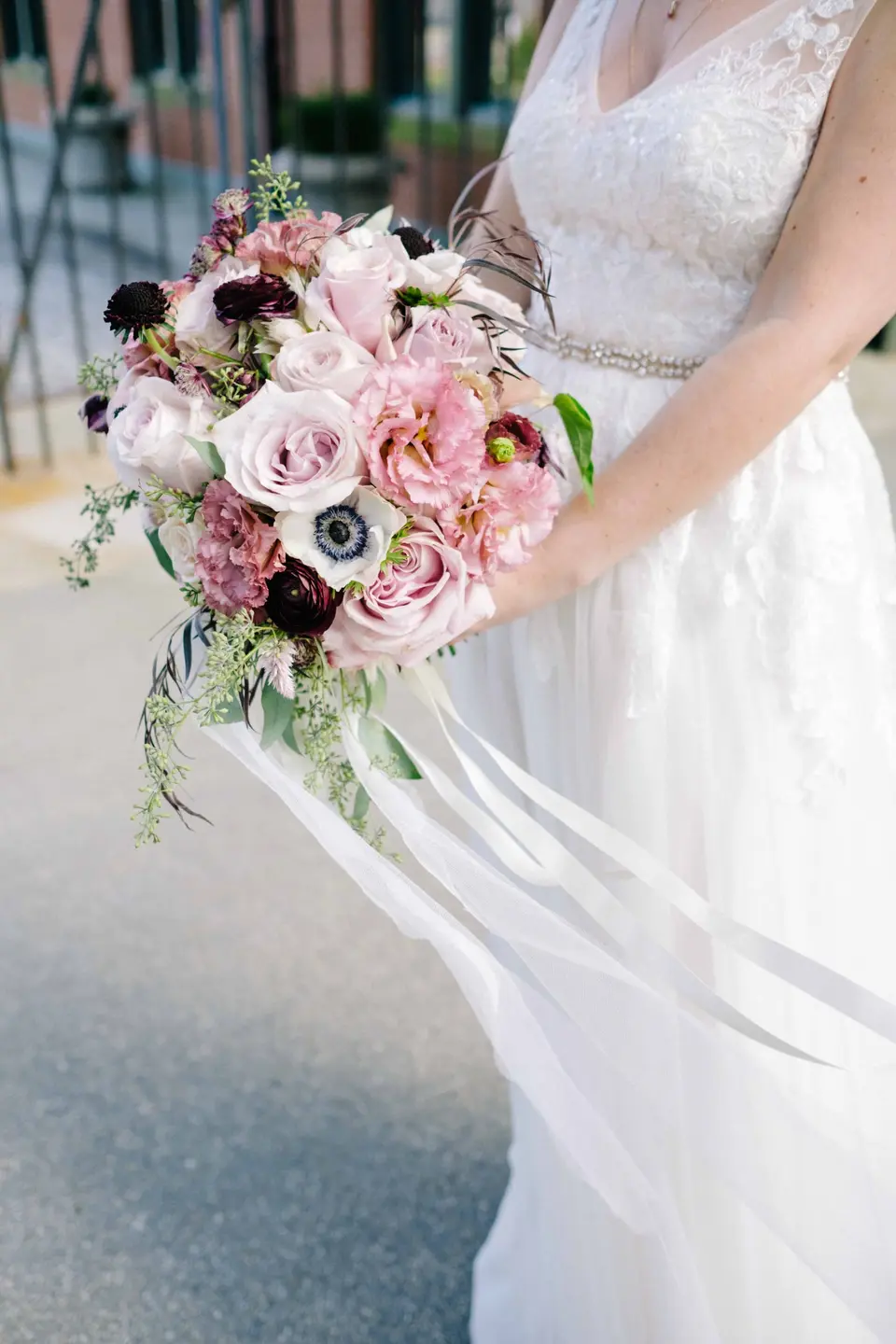 The bride holds a loose bouquet of pink roses, anemones and deep plum blooms, silk ribbons trailing in the wind.