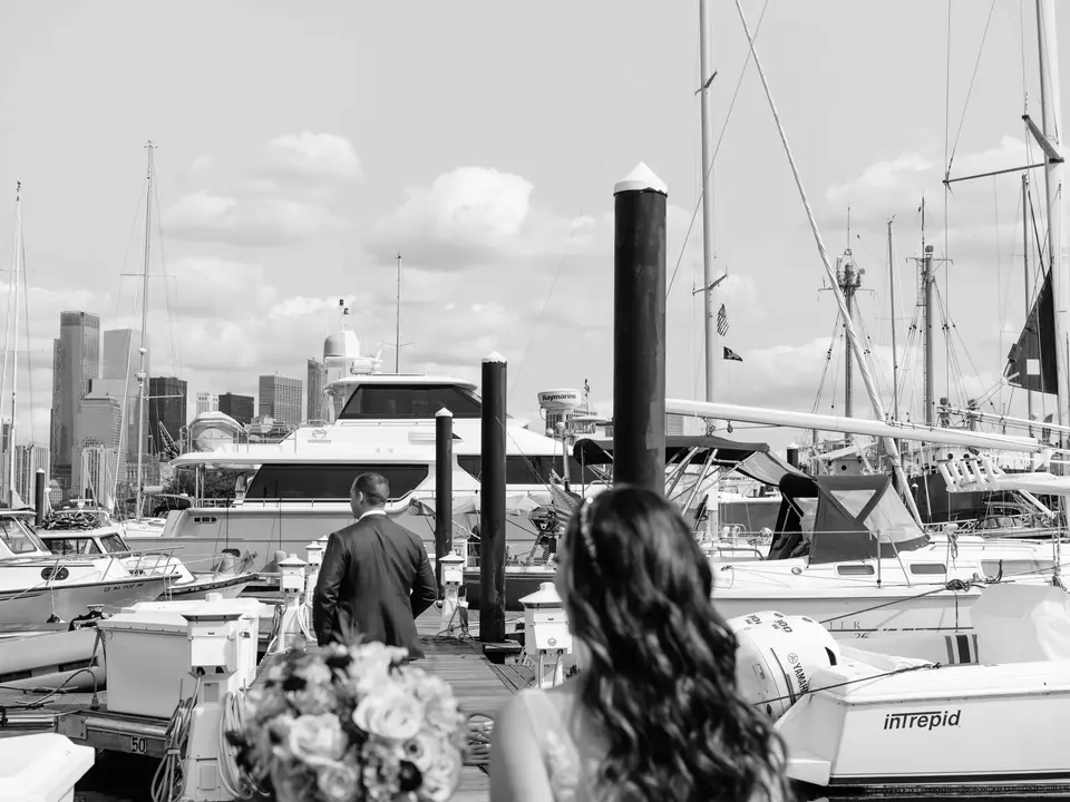 The bride and groom walk out along a marina dock, sailboats and the Jersey City skyline ahead. Black and white.