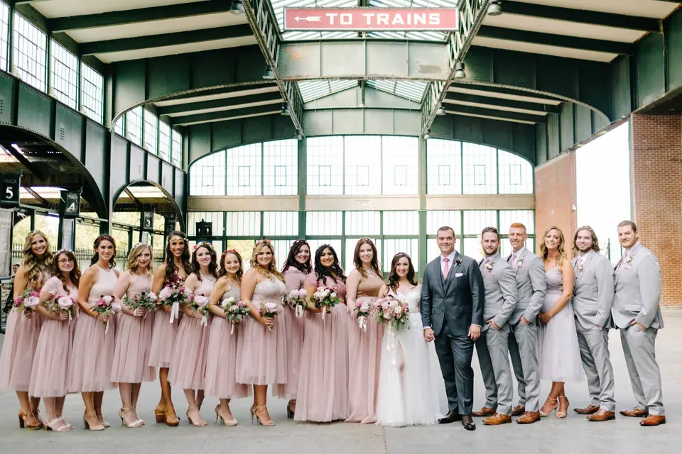 The wedding party lined up in the old train terminal beneath a To Trains sign, the bridesmaids in blush and the groomsmen in grey.