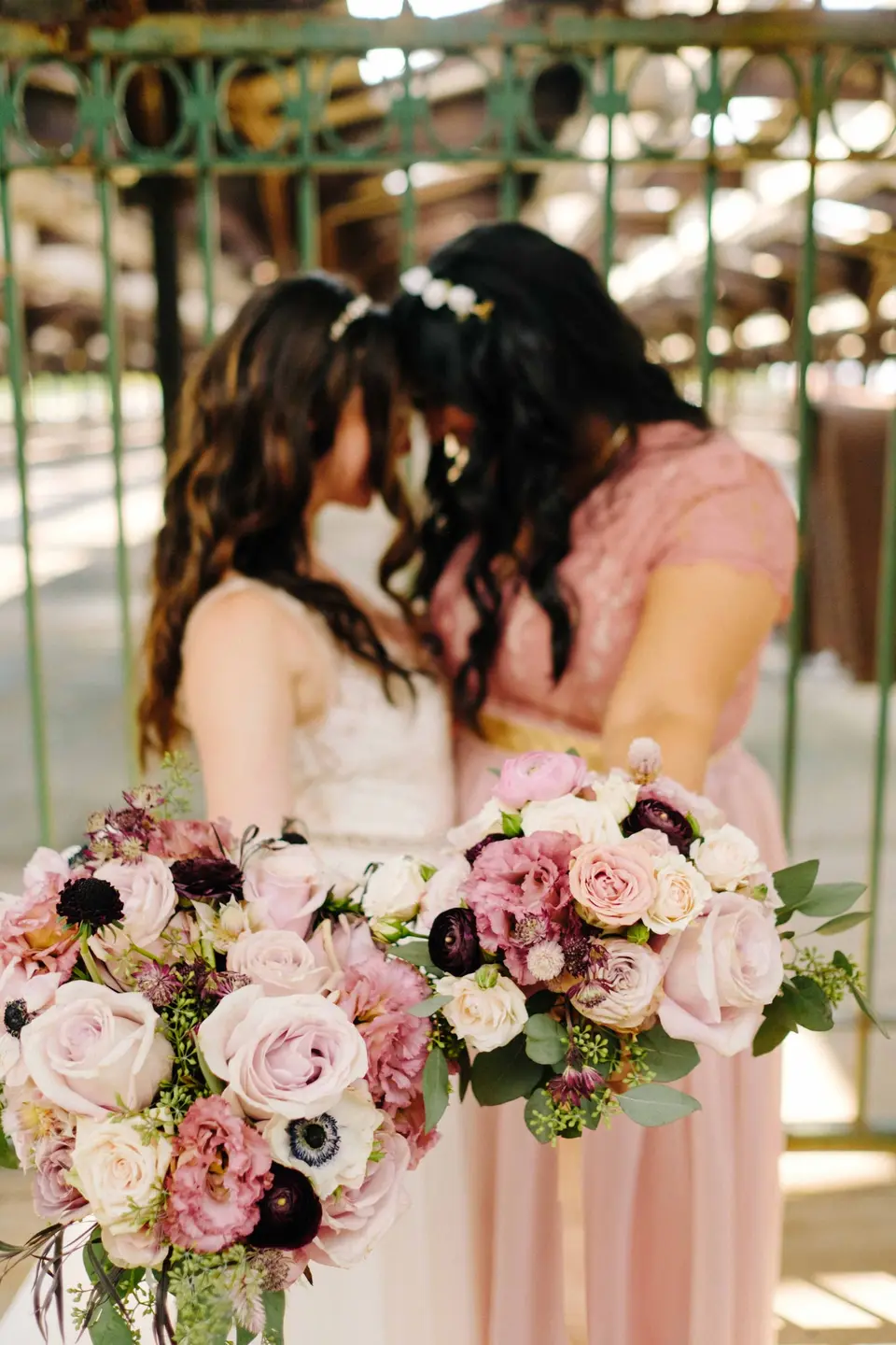 The bride and a bridesmaid in blush touch foreheads, their bouquets of pink roses held low.