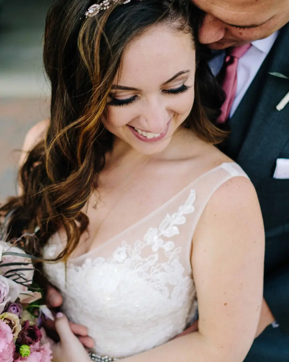 The groom holds the bride close from behind as she smiles down at her bouquet.