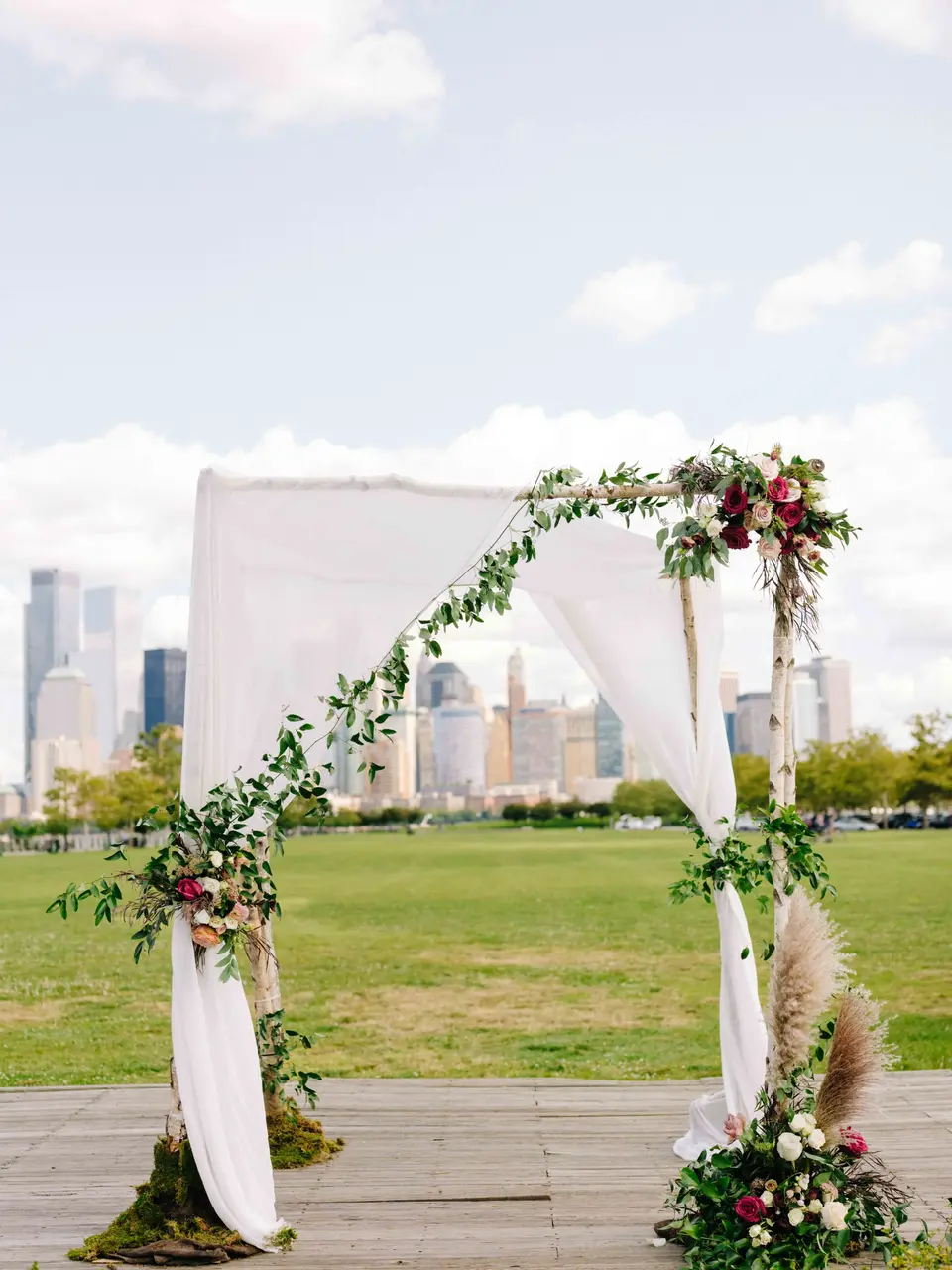 A draped wooden arch trimmed with greenery, roses and pampas grass on the lawn, Lower Manhattan beyond.