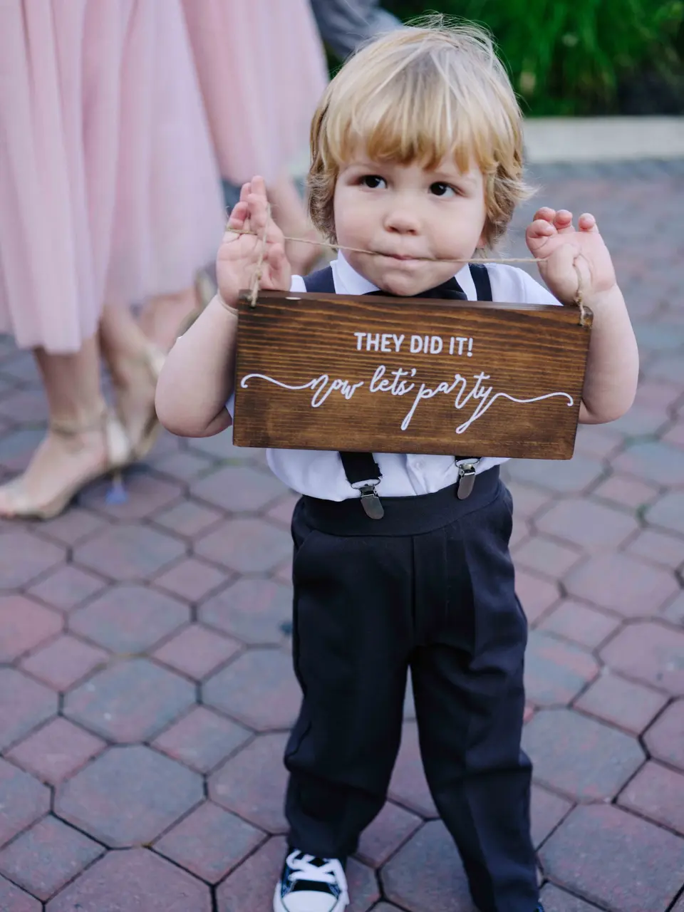 A little boy in suspenders holds a wooden sign reading They did it, now let's party.