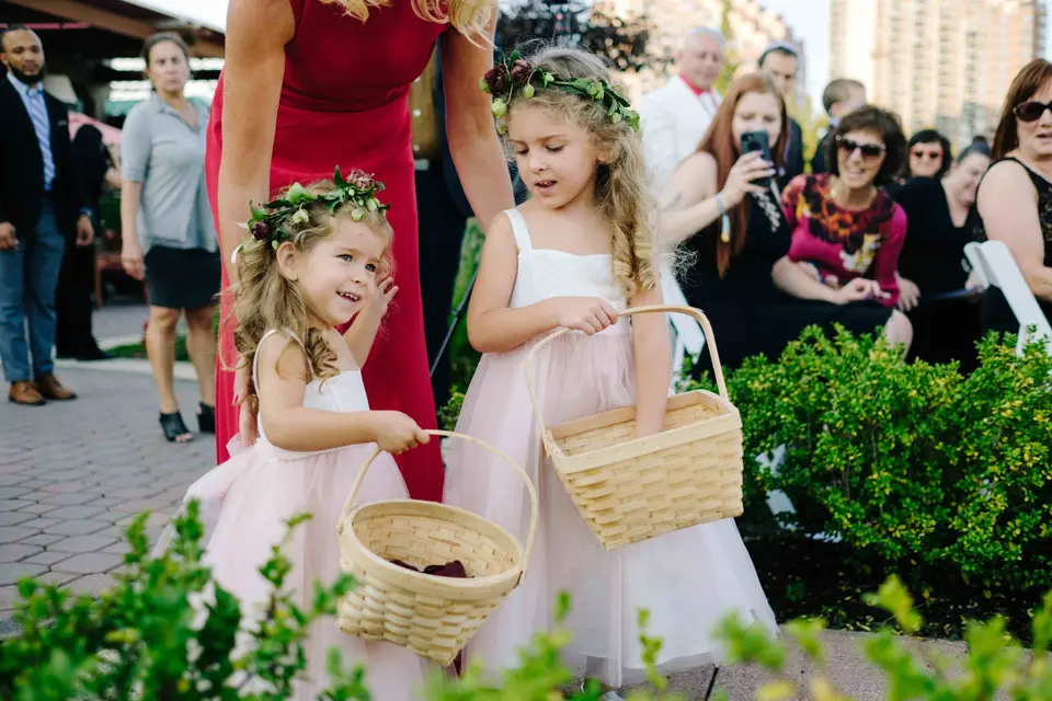 Two flower girls in floral crowns scatter petals from woven baskets as guests watch.