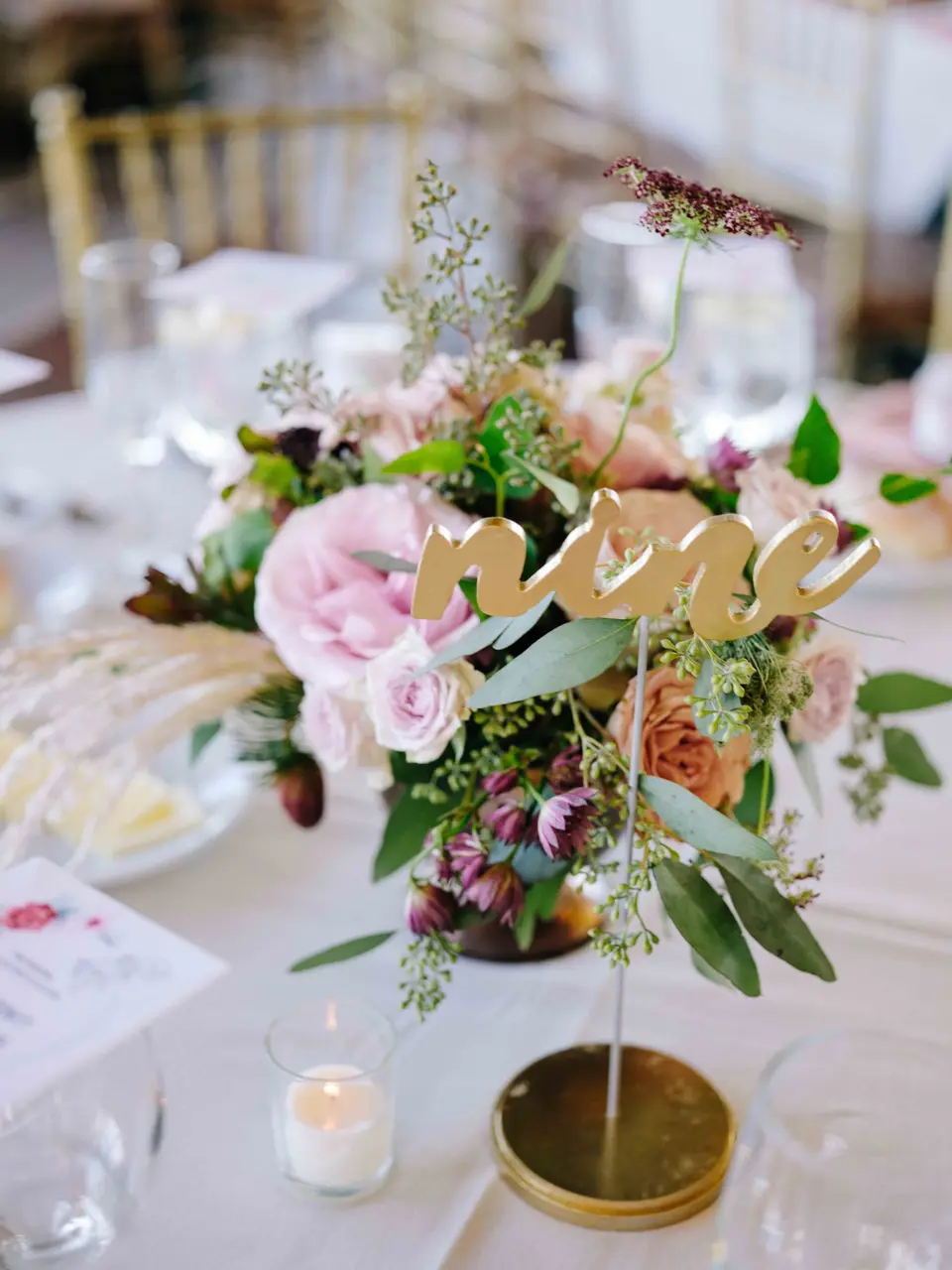 A centerpiece of blush roses and greenery with a gold table number, a votive candle beside it.