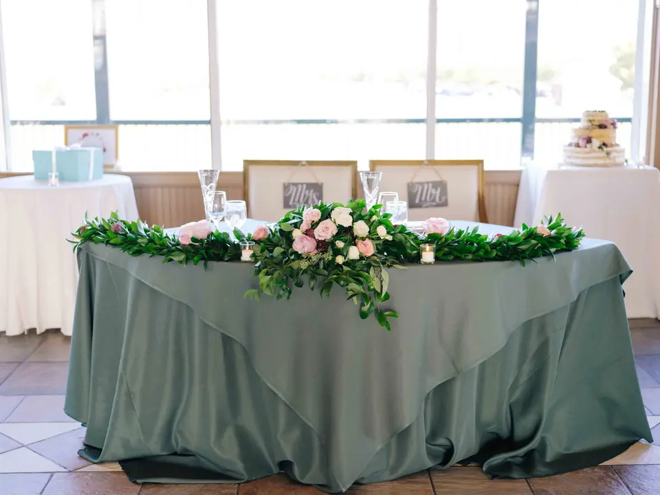 The sweetheart table in sage green, garlanded with greenery and pink roses, the cake on a side table by the windows.