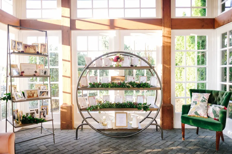 A round shelf of framed photographs and greenery in a sunlit window bay, a green velvet chair beside it.