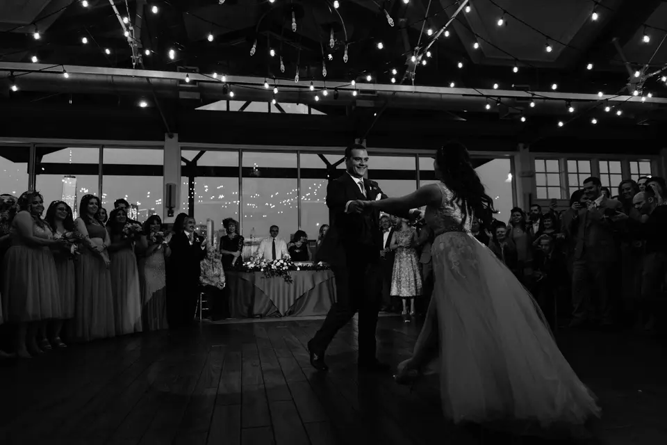 The first dance beneath strings of lights, guests watching from the edges of the floor. Black and white.