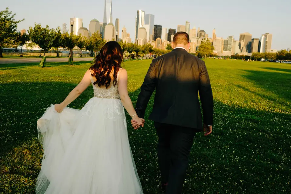 The bride and groom walk hand in hand across the lawn toward the Lower Manhattan skyline.
