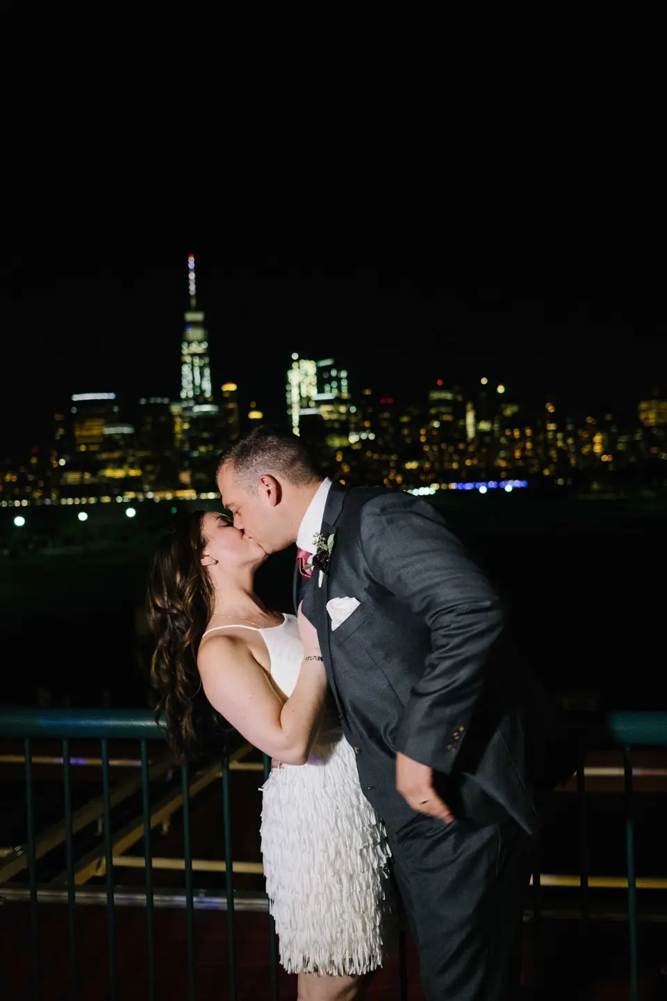 At night, the groom dips the bride for a kiss, One World Trade Center lit up across the water.