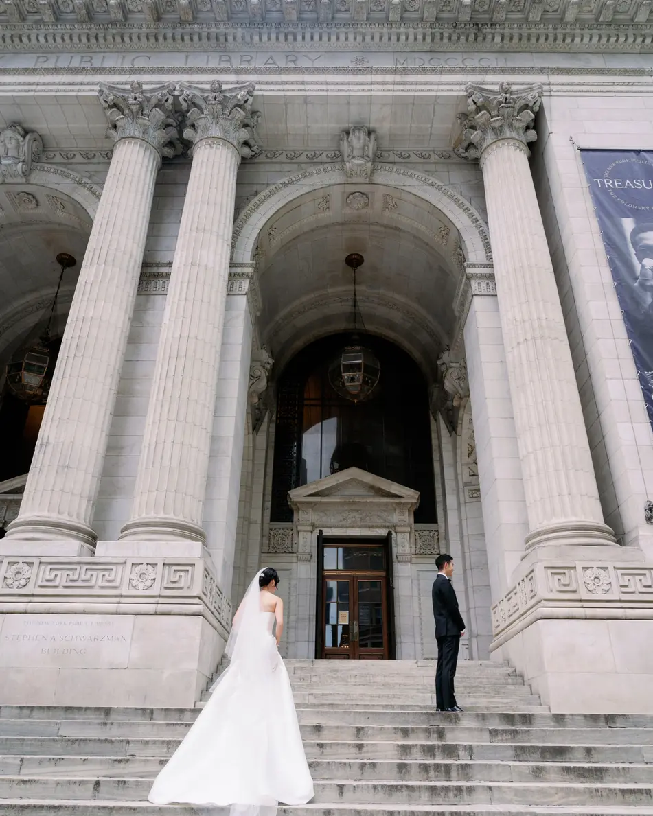 The bride and groom stand on the wide stone steps of the New York Public Library on Fifth Avenue, between its great columns.