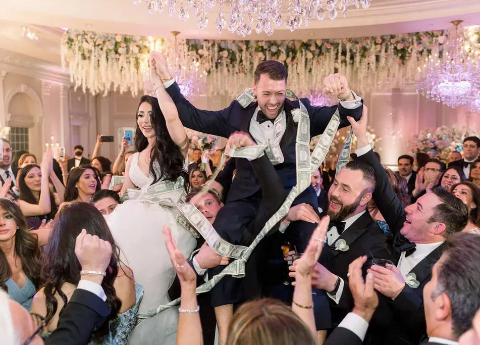 The groom is lifted high above a cheering crowd on the dance floor beneath chandeliers and hanging white flowers.