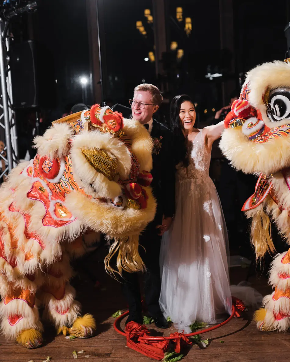 Two lion dancers in red, gold and cream rear up on either side of the couple beneath stage lights at Blue Hill at Stone Barns, guests gathered behind them.