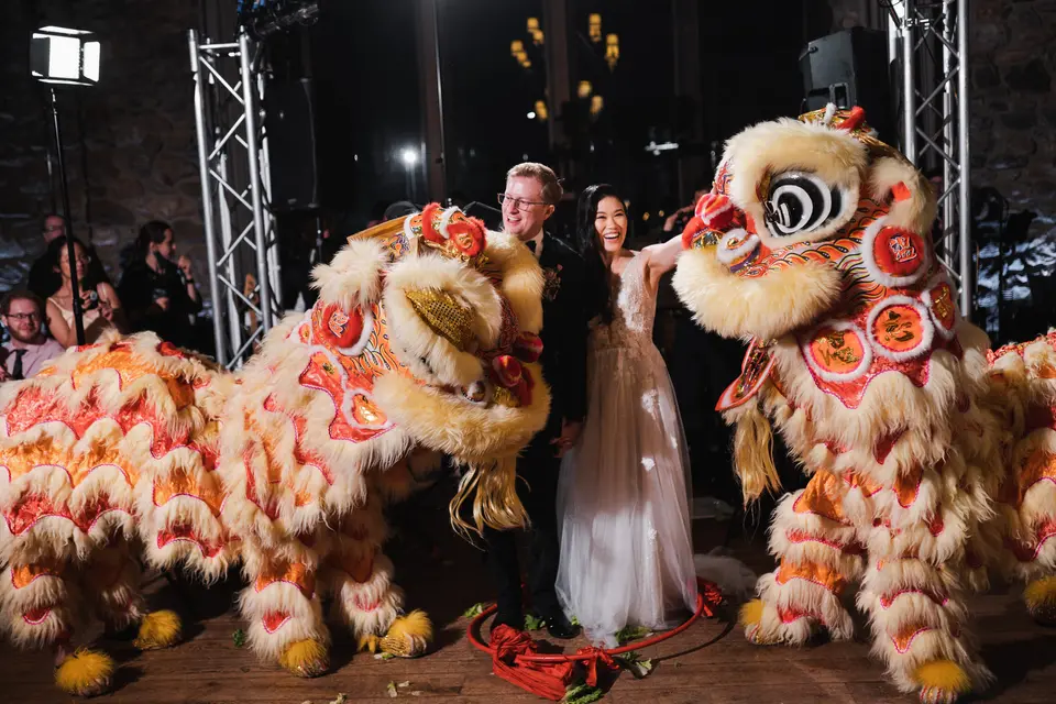 Two lion dancers in red, gold and cream rear up on either side of the couple beneath stage lights at Blue Hill at Stone Barns, guests gathered behind them.