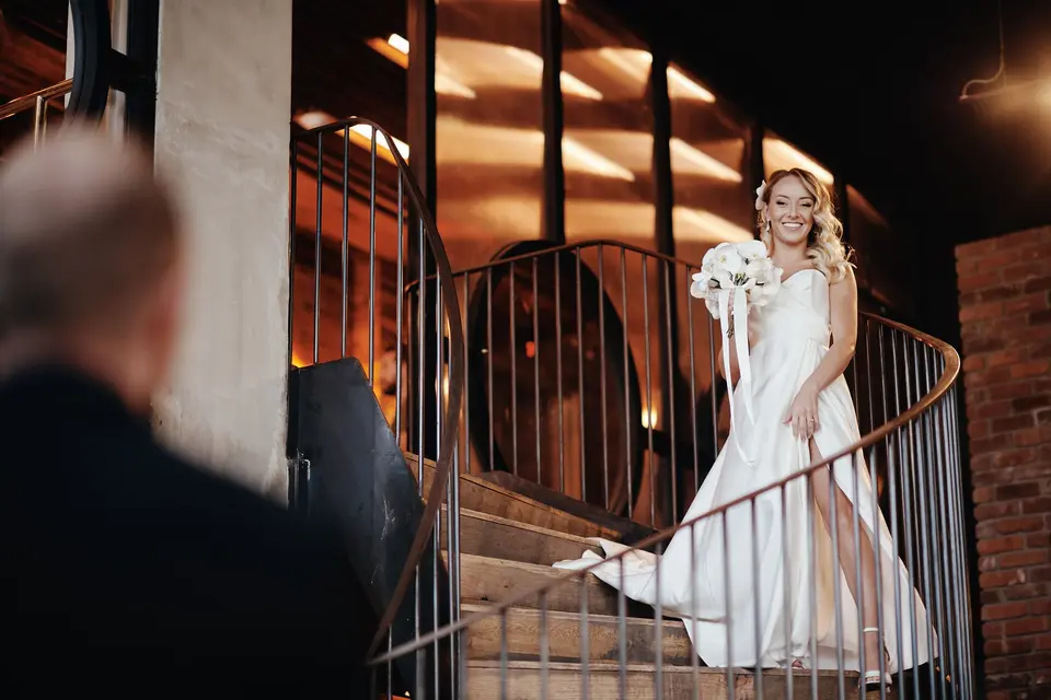 The bride comes down the spiral staircase holding white orchids, the groom watching from below.