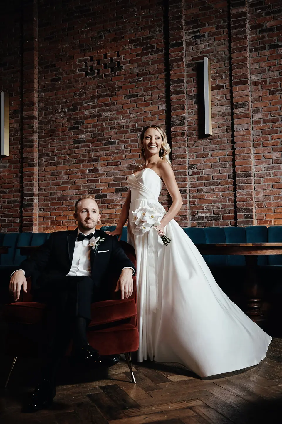 Against a brick wall, the groom sits in a velvet chair and the bride stands beside him in her gown.