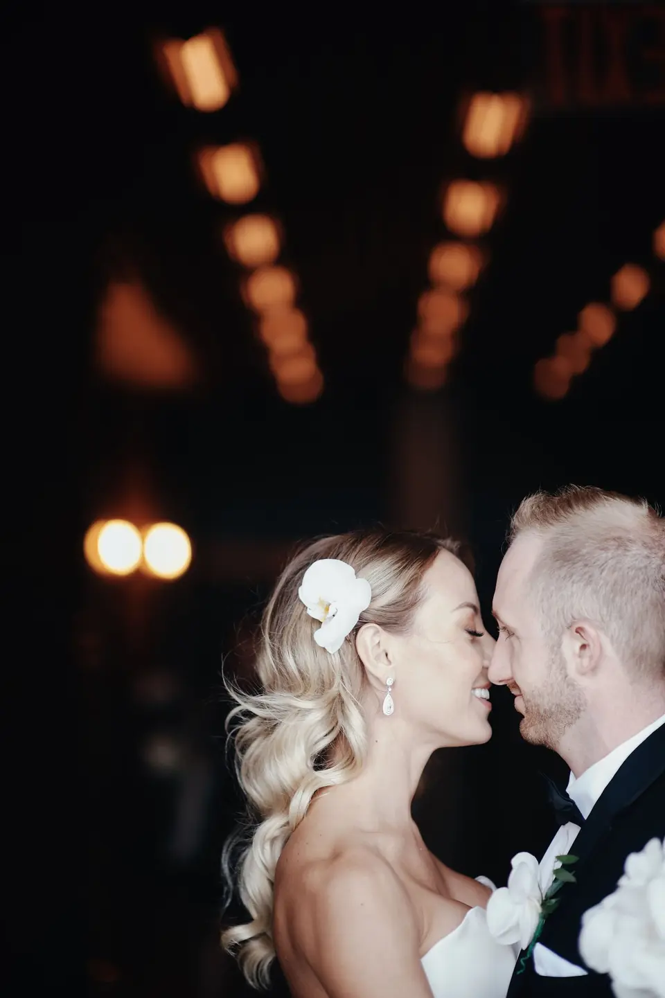 The bride and groom touch noses beneath warm lights, an orchid in her hair.