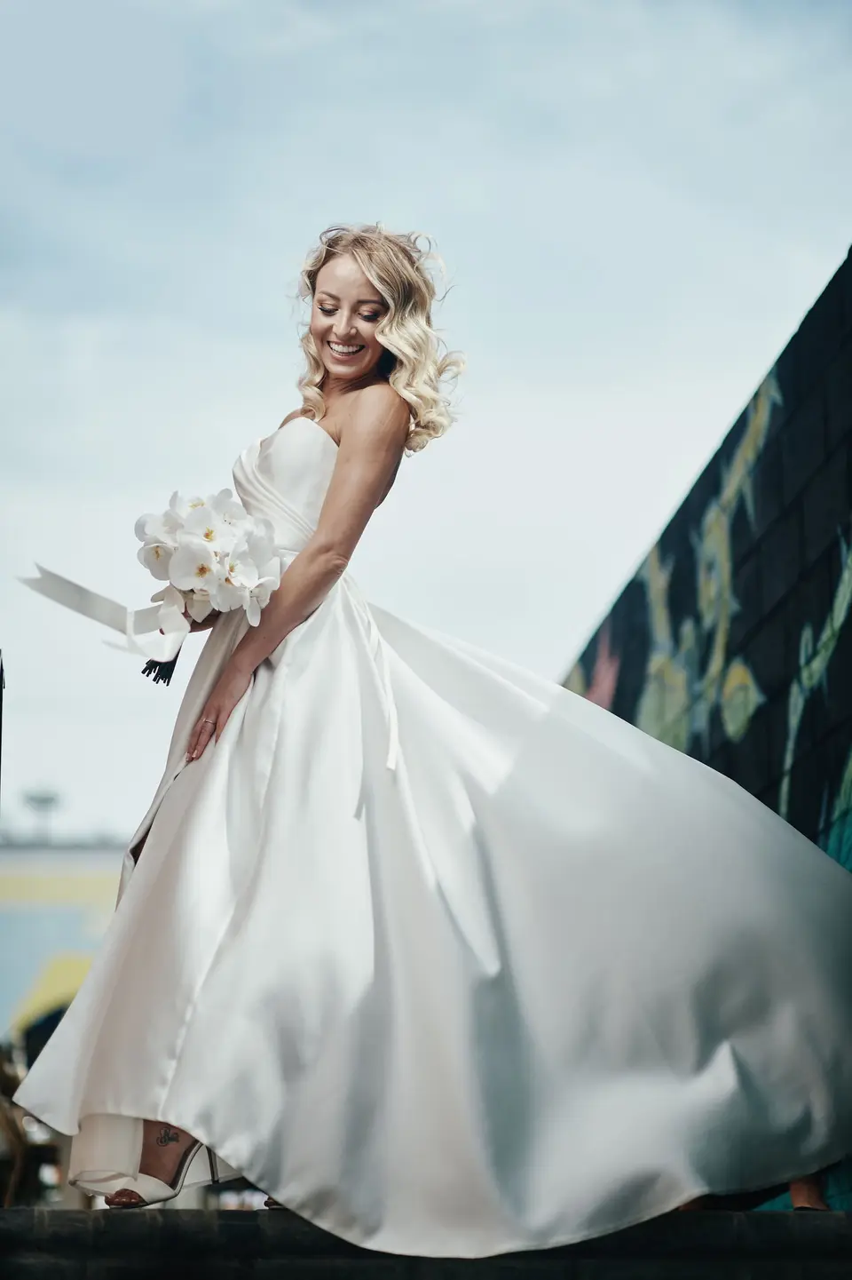 The bride twirls in her satin gown against the sky, orchids in her hand.