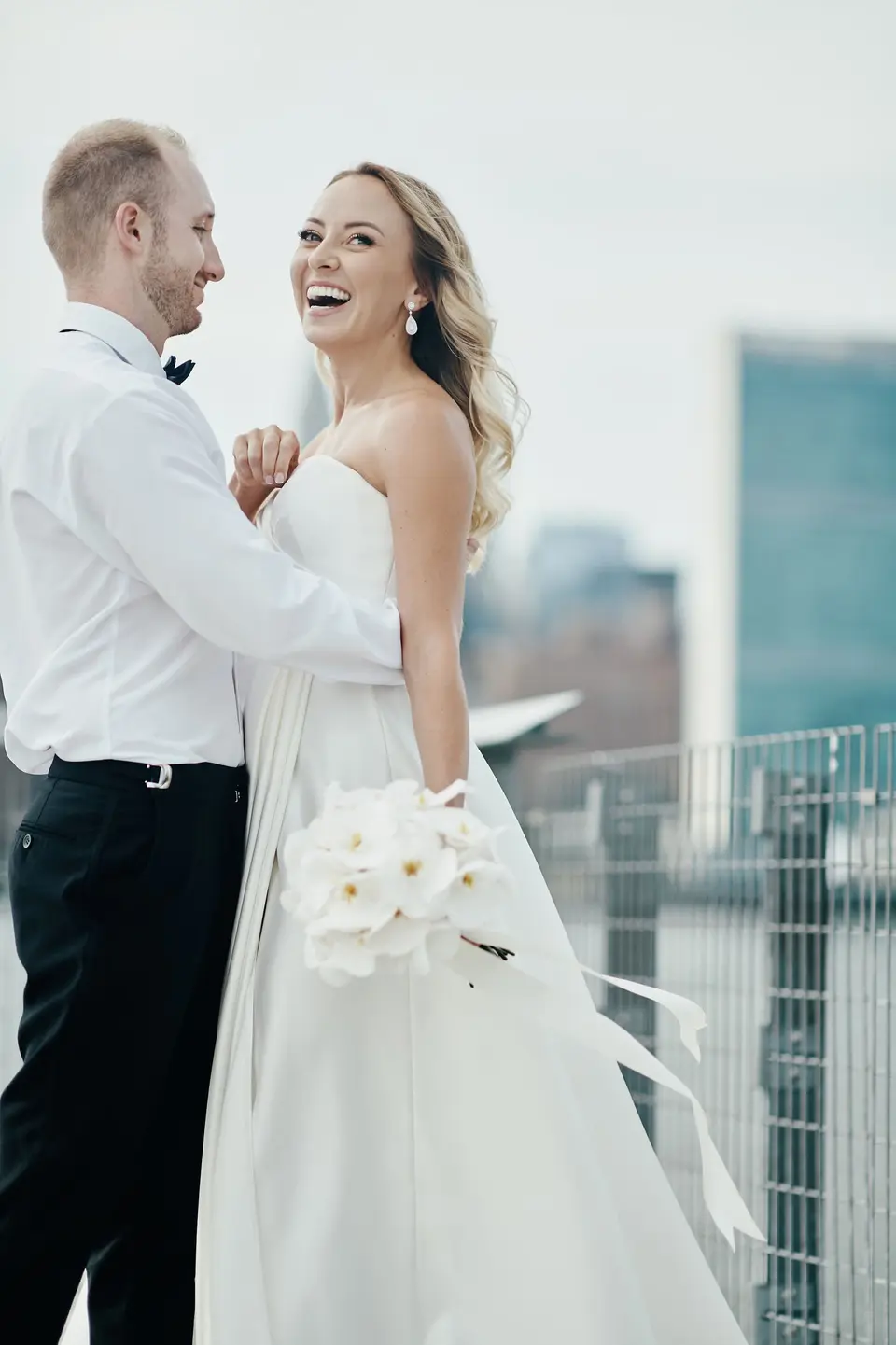 The groom in shirtsleeves holds the bride as she laughs, the East River and Midtown behind them.