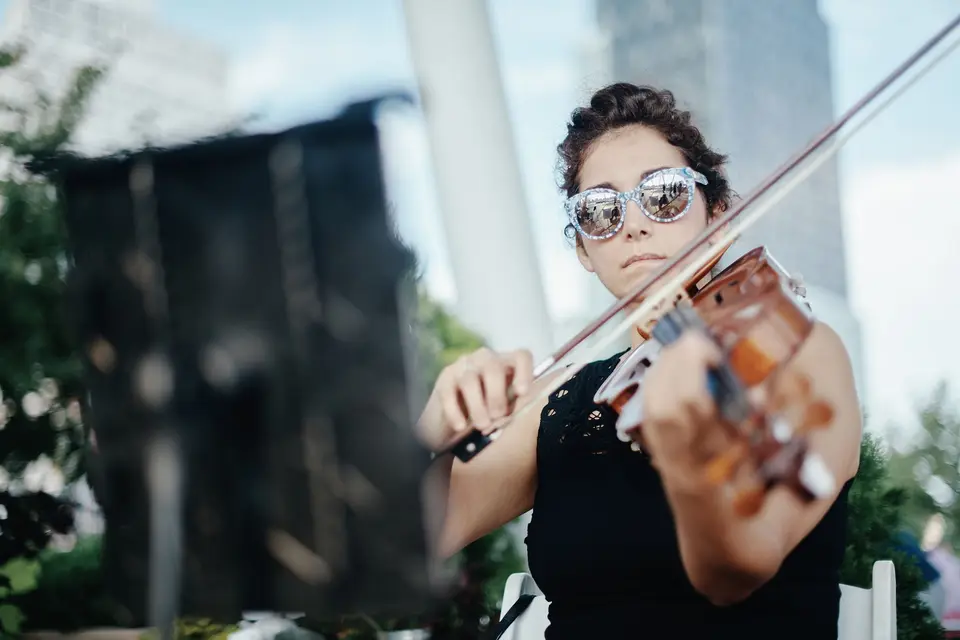 A violinist in mirrored sunglasses plays outdoors, a tower behind her.