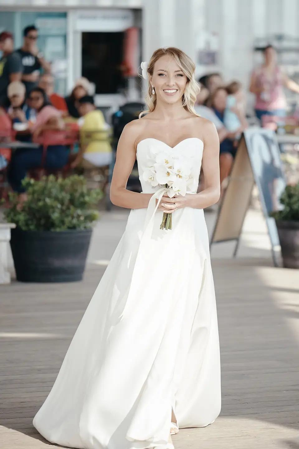 The bride walks along the waterfront promenade holding her orchids, people at café tables behind her.