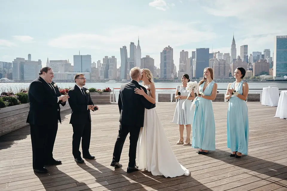 On the waterfront deck, the groom embraces the bride as family and bridesmaids in pale blue look on, the Manhattan skyline behind.