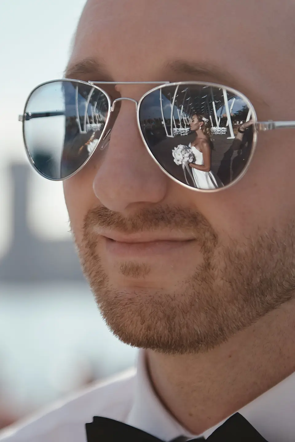 The bride and her bouquet reflected in the groom's aviator sunglasses as he smiles.