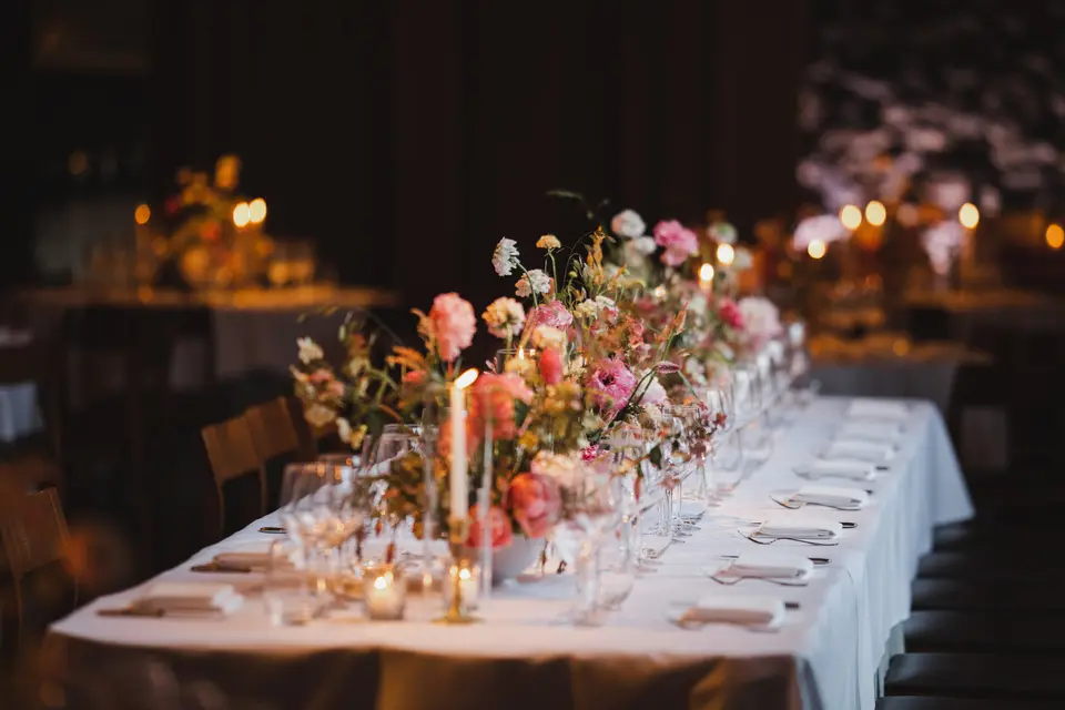 A long table set with candles and loose arrangements of pink and peach flowers glows in a dark room.