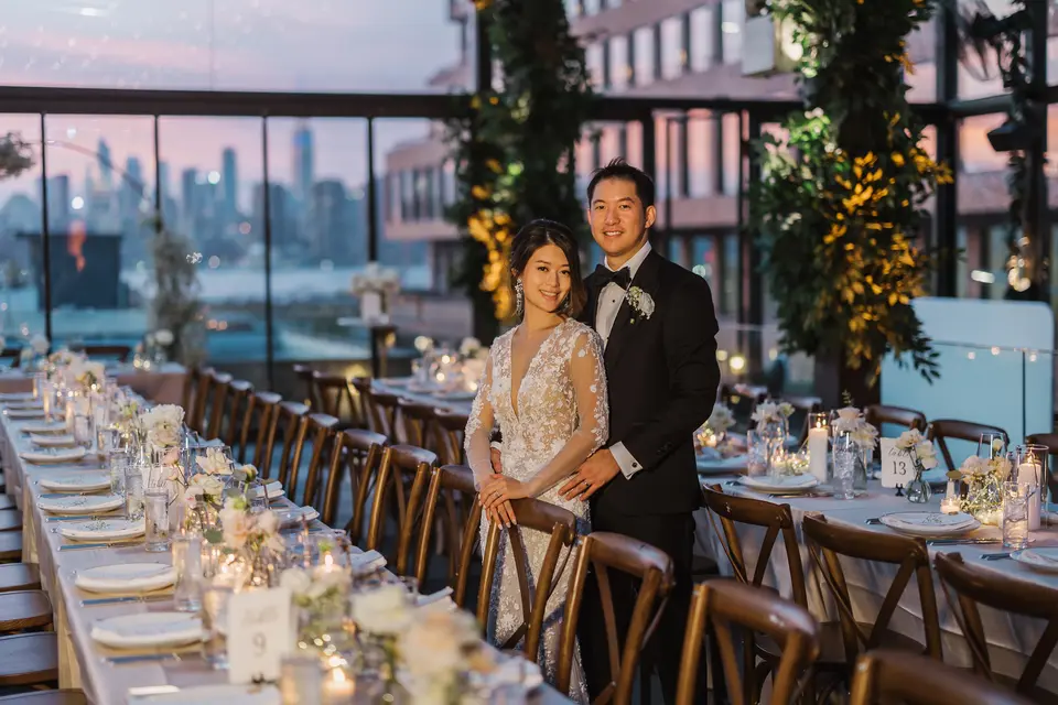 The couple stand at the end of long wooden tables set with candles and white flowers, the skyline turning pink at dusk behind the glass.
