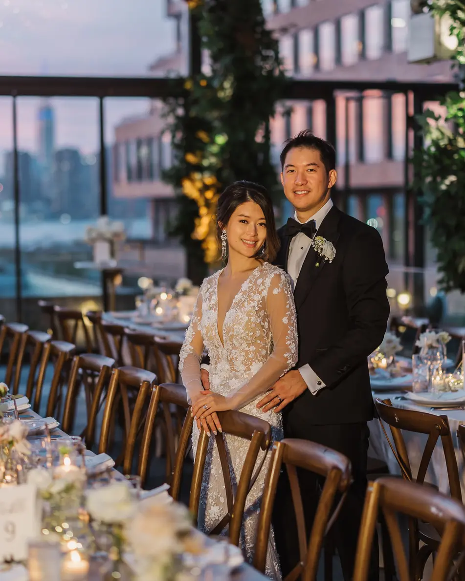 The couple stand at the end of long wooden tables set with candles and white flowers, the skyline turning pink at dusk behind the glass.