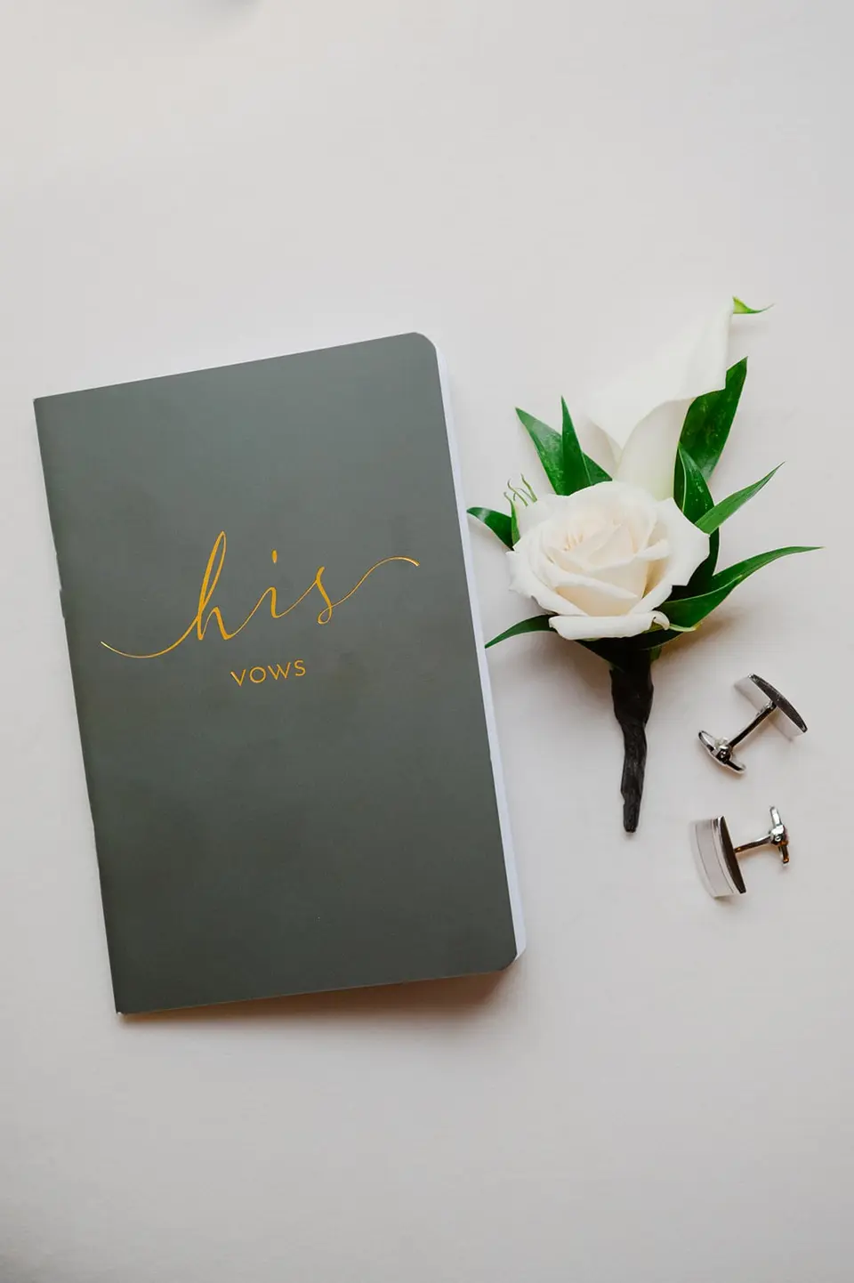 A green vow book lettered His vows in gold, beside a white rose boutonniere and cufflinks.